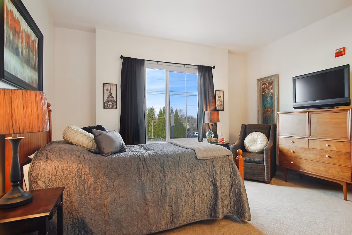 Well-lit furnished bedroom with a bed, nightstand and lamp, an armchair, a dresser with a TV, and a large window with dark curtains.