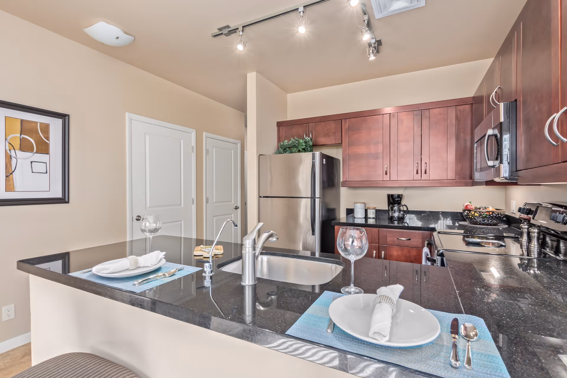 Modern kitchen with dark granite countertops and wooden cabinets. The kitchen features a stainless steel refrigerator, microwave, stove, and a coffee maker. There is a kitchen island with a sink and two place settings, each with a plate, napkin, utensils, and a wine glass. Two white doors are visible in the background along with a framed abstract artwork on the wall.