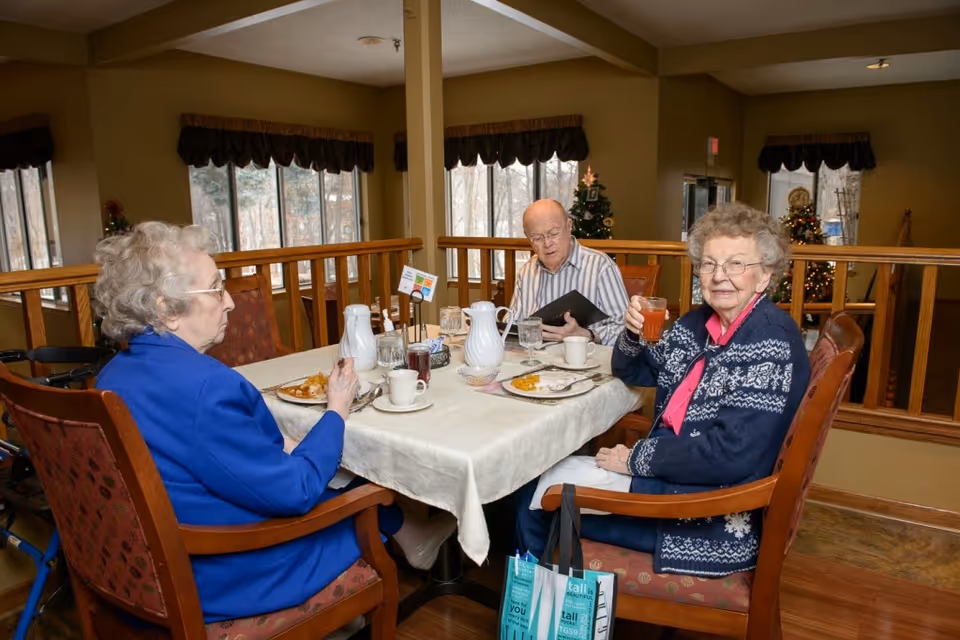 Three elderly people sitting around a table in a dining area. Two women and one man are seated, with plates of food and drinks on the table. One woman is holding up a glass and smiling at the camera. The room has large windows with curtains and Christmas decorations in the background.