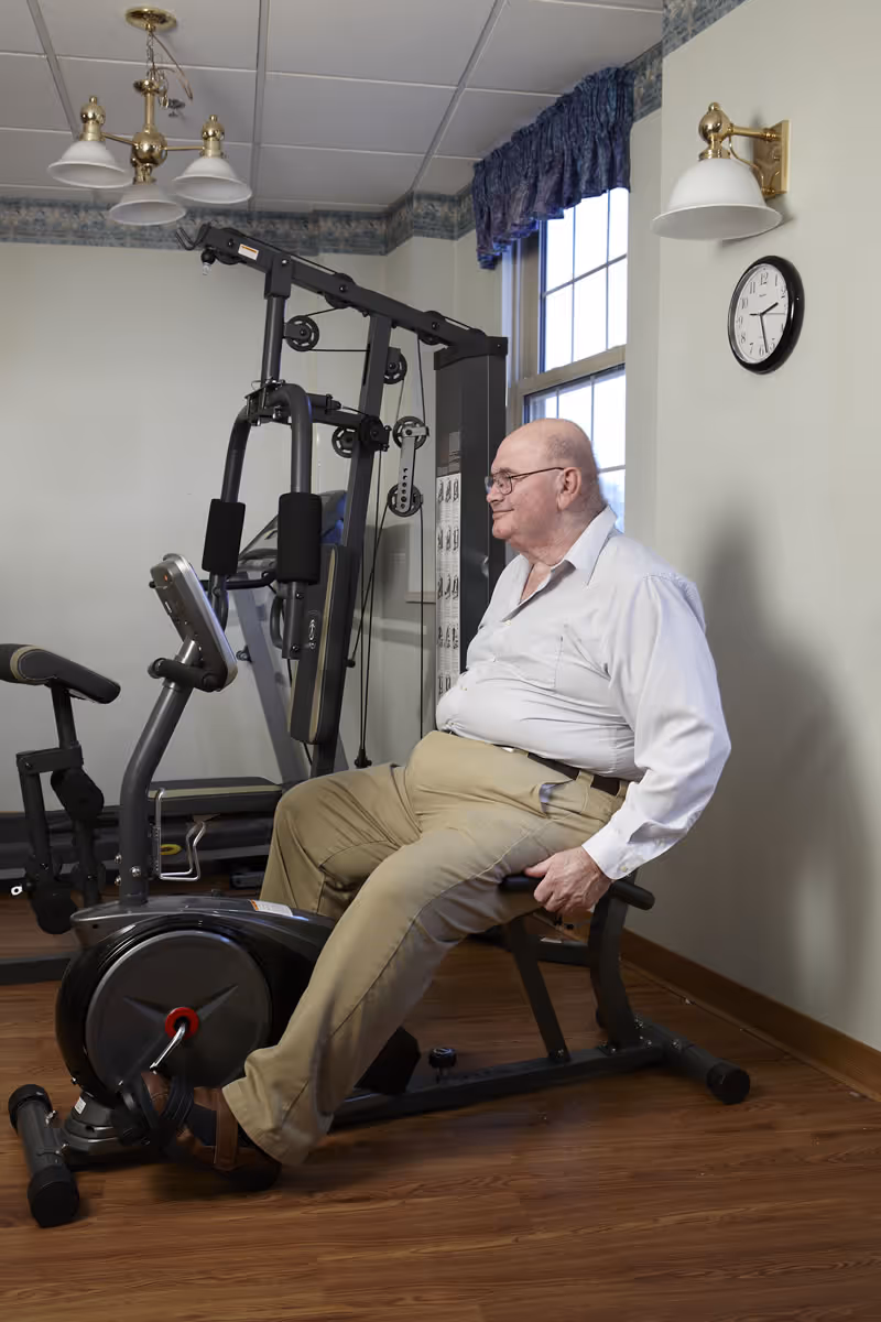An elderly man wearing glasses, a white shirt, and beige pants is seated on a stationary exercise bike in a room with wooden flooring. Behind him, there is a multi-functional gym machine. The room has a window with blue curtains, a wall clock showing the time as 3:40, and ceiling and wall-mounted light fixtures.