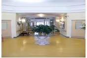 Spacious nursing home lobby with a central decorative plant on a pedestal, flanked by doorways and seating areas.