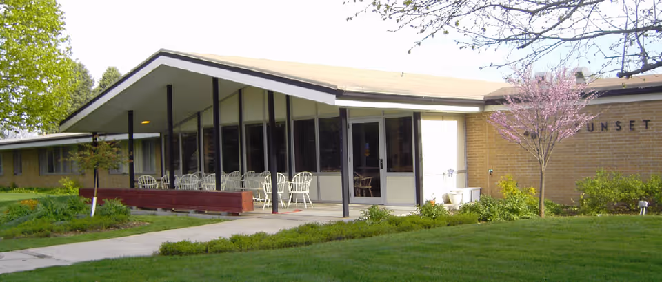 Exterior view of a single-story building with large windows and a covered patio area furnished with white chairs and tables. The building is surrounded by green grass, shrubs, and a small tree with pink blossoms. The sky is clear and the scene appears to be during daytime.