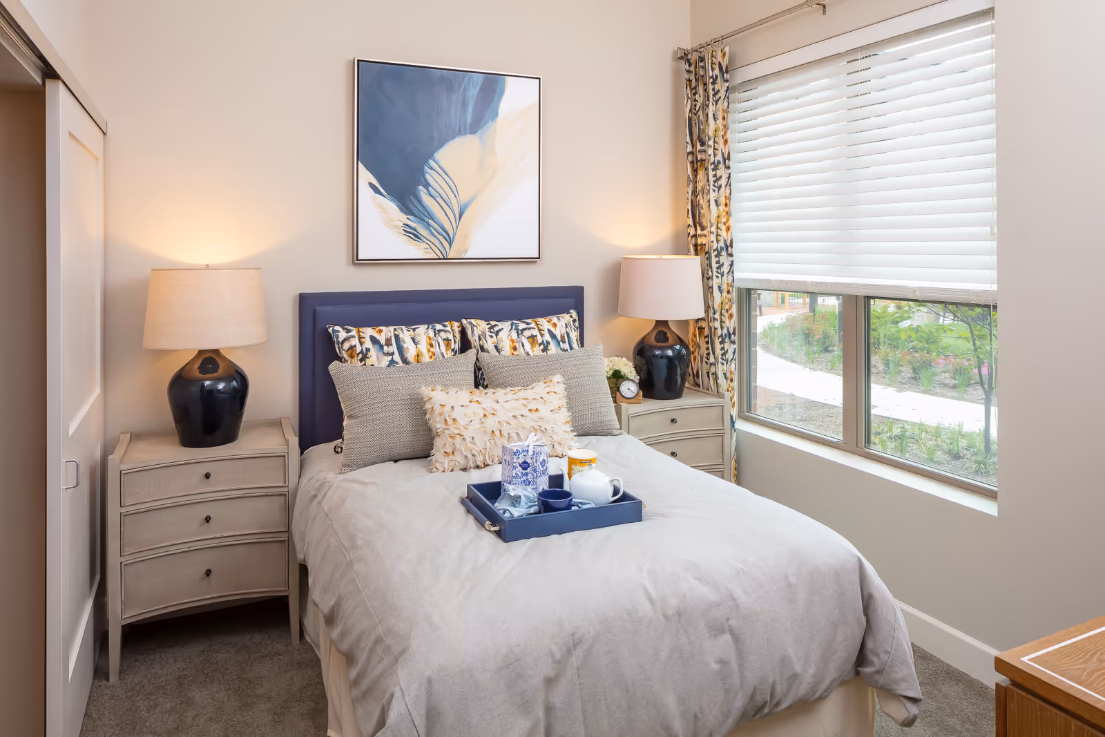 Sunlit bedroom with a made bed topped by decorative pillows and a tray, flanked by matching nightstands and lamps beneath framed artwork and a large window.