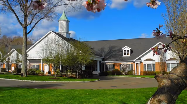 Exterior view of a single-story senior living facility building with a white facade, brick accents, and a small cupola on the roof. The building is surrounded by green grass, trees with budding leaves, and flowering branches in the foreground under a blue sky with some clouds.