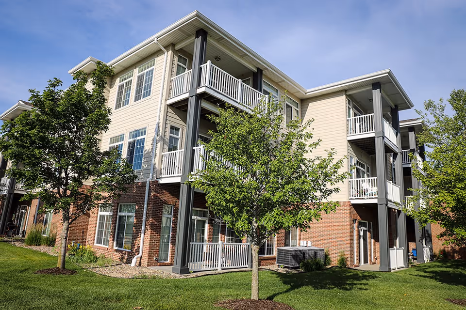 Three-story senior living building with balconies, brick and light-colored siding exterior, and a grassy lawn with trees.