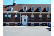 Exterior front view of a two-story brick building with a white entrance door, multiple windows, and a brown shingled roof with dormer windows. The building is situated next to a paved parking area under a clear blue sky.