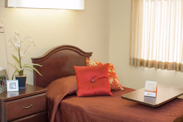A bedroom with a single wooden bed covered with a brown quilt and two decorative pillows, one red and one with a red and beige pattern. Next to the bed is a wooden nightstand with a potted orchid plant and a framed photo. A window with beige curtains allows natural light into the room. A small wooden tray with a welcome card is placed on the bed.