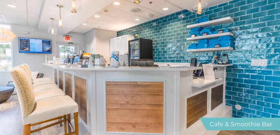 Interior view of a cafe and smoothie bar with a white counter featuring wood paneling, several cushioned chairs lined up along the counter, teal subway tile backsplash, shelves with neatly stacked blue towels, and digital menu screens on the wall.