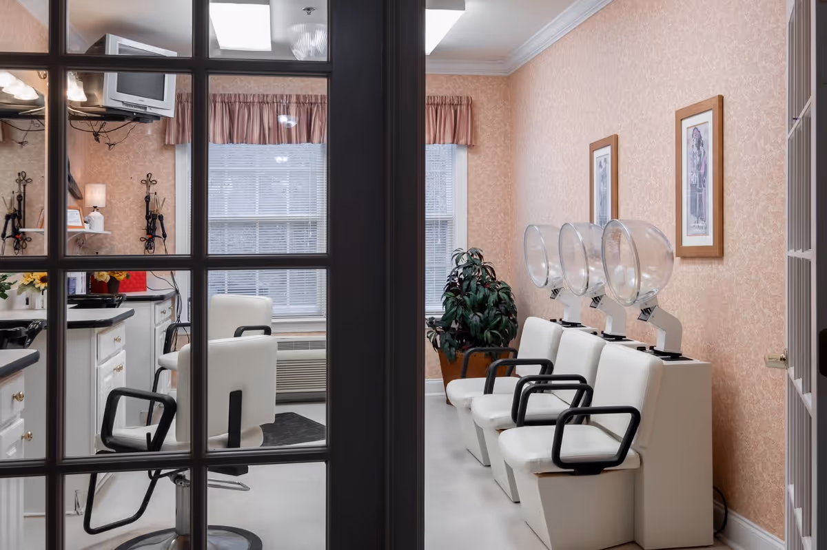 Interior view of a salon area in a senior living facility with white salon chairs and hair drying stations along a peach-colored wall. The room has large windows with blinds and pink valances, a potted plant, framed artwork on the wall, and a small TV mounted in the corner. The photo is taken through glass-paneled double doors.