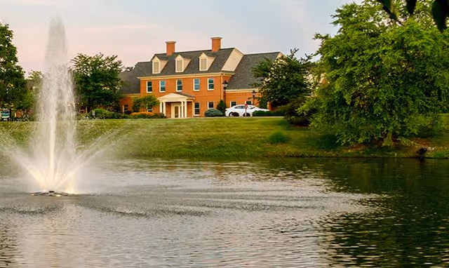 A large brick building with a gray roof and multiple chimneys sits behind a grassy area with trees. In the foreground, there is a pond with a water fountain spraying water upwards.
