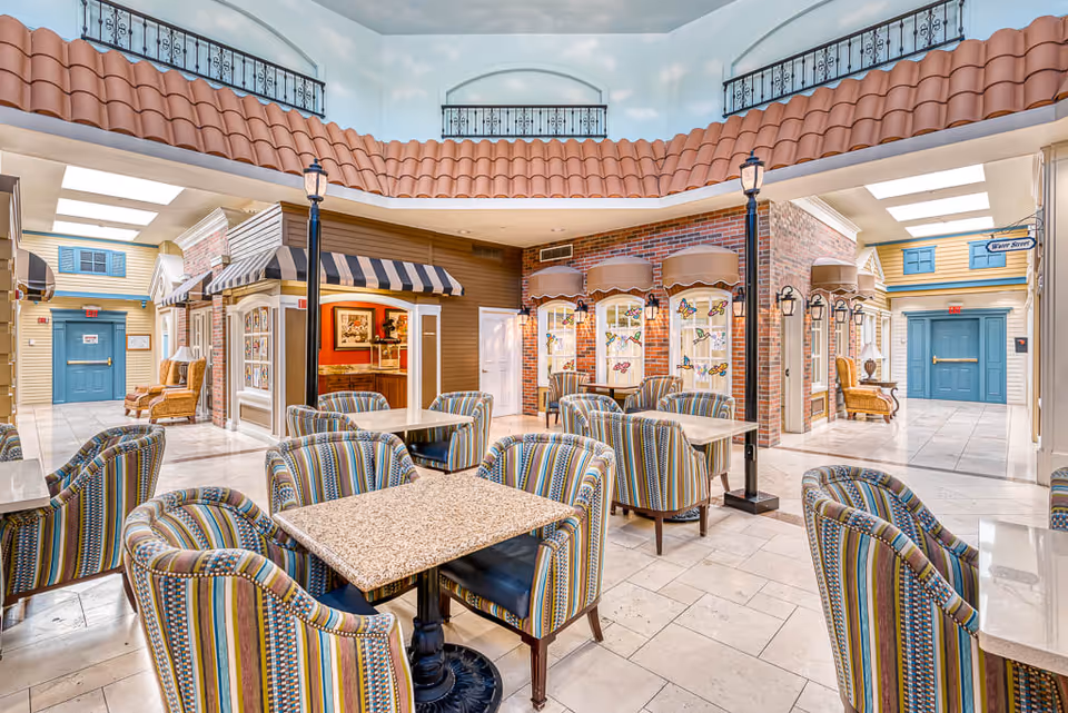 Bright and colorful indoor common area with multiple tables and striped upholstered chairs arranged for seating. The space features a high ceiling with a sky mural and decorative street lamps. Surrounding walls are designed to resemble a quaint small town with storefronts, awnings, and windows decorated with butterfly decals. Blue double doors and comfortable armchairs are visible in the background.