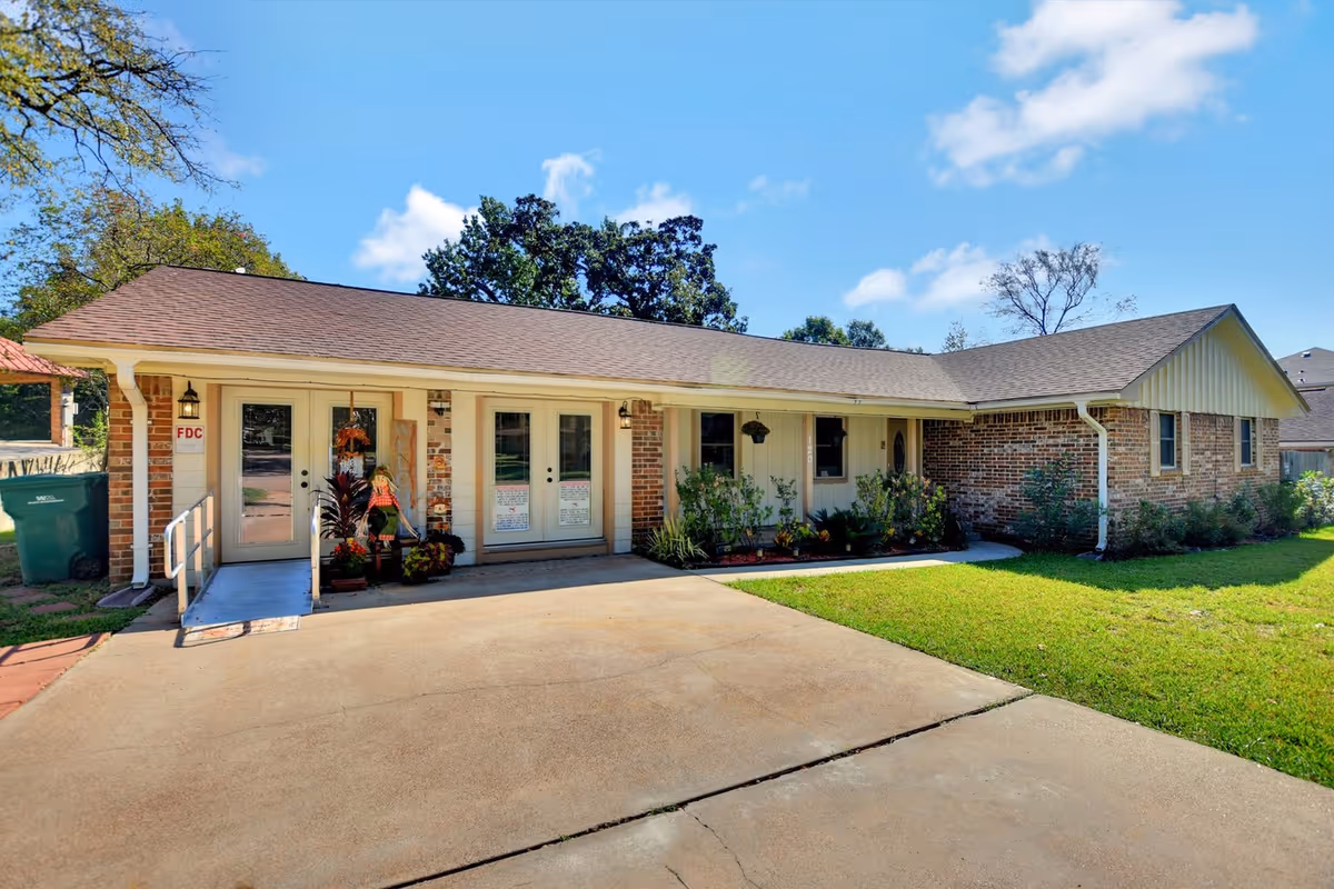 Single-story brick building with a brown shingled roof, featuring two glass doors with white frames and a small ramp leading to the entrance. The building is surrounded by green grass and some plants near the entrance. The sky is clear with a few clouds.