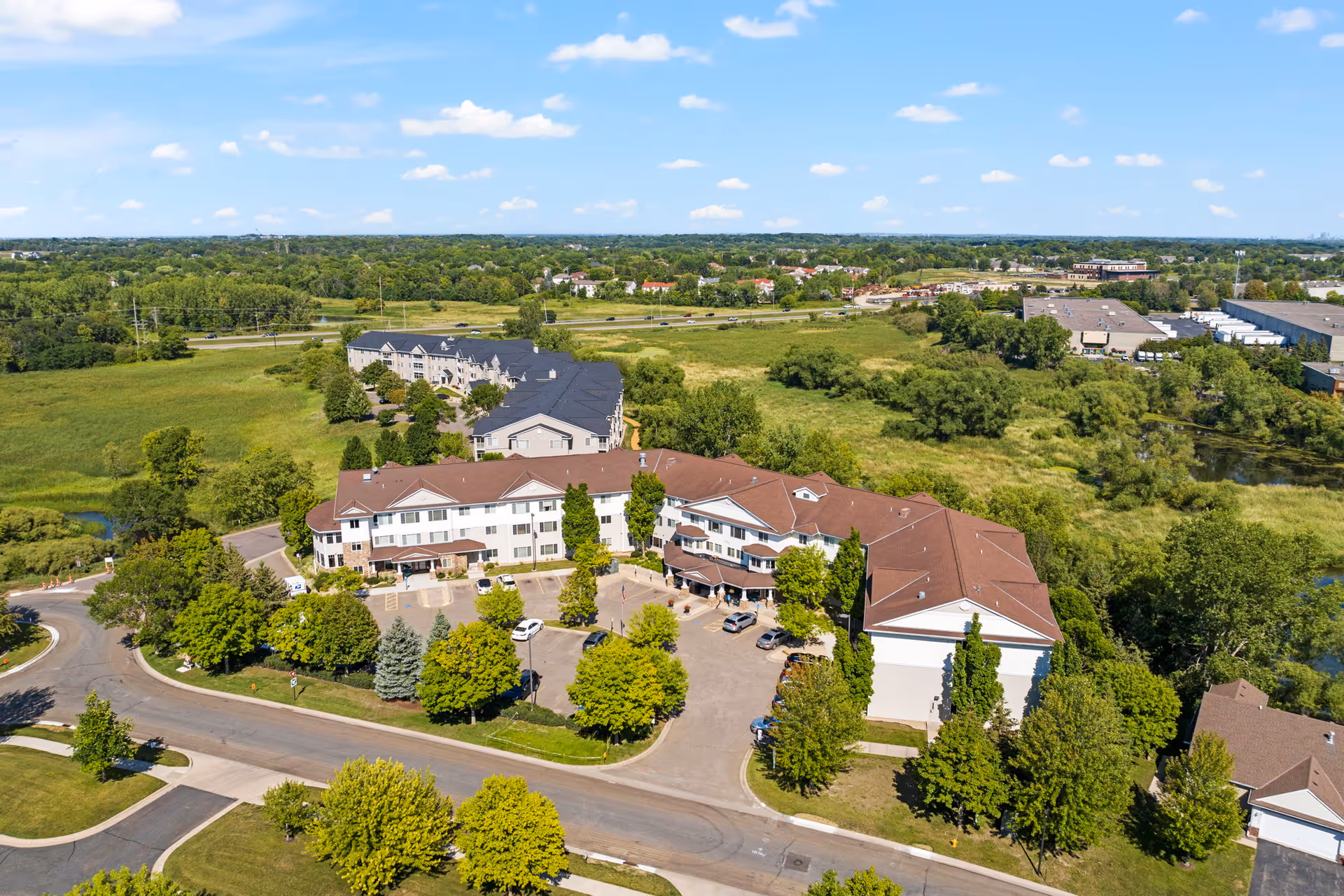 Aerial view of a large assisted living and memory care facility surrounded by greenery and trees, with a parking lot and several cars in front. The building has multiple wings with brown and black roofs, situated in a suburban area with open fields and other buildings in the distance under a blue sky with scattered clouds.