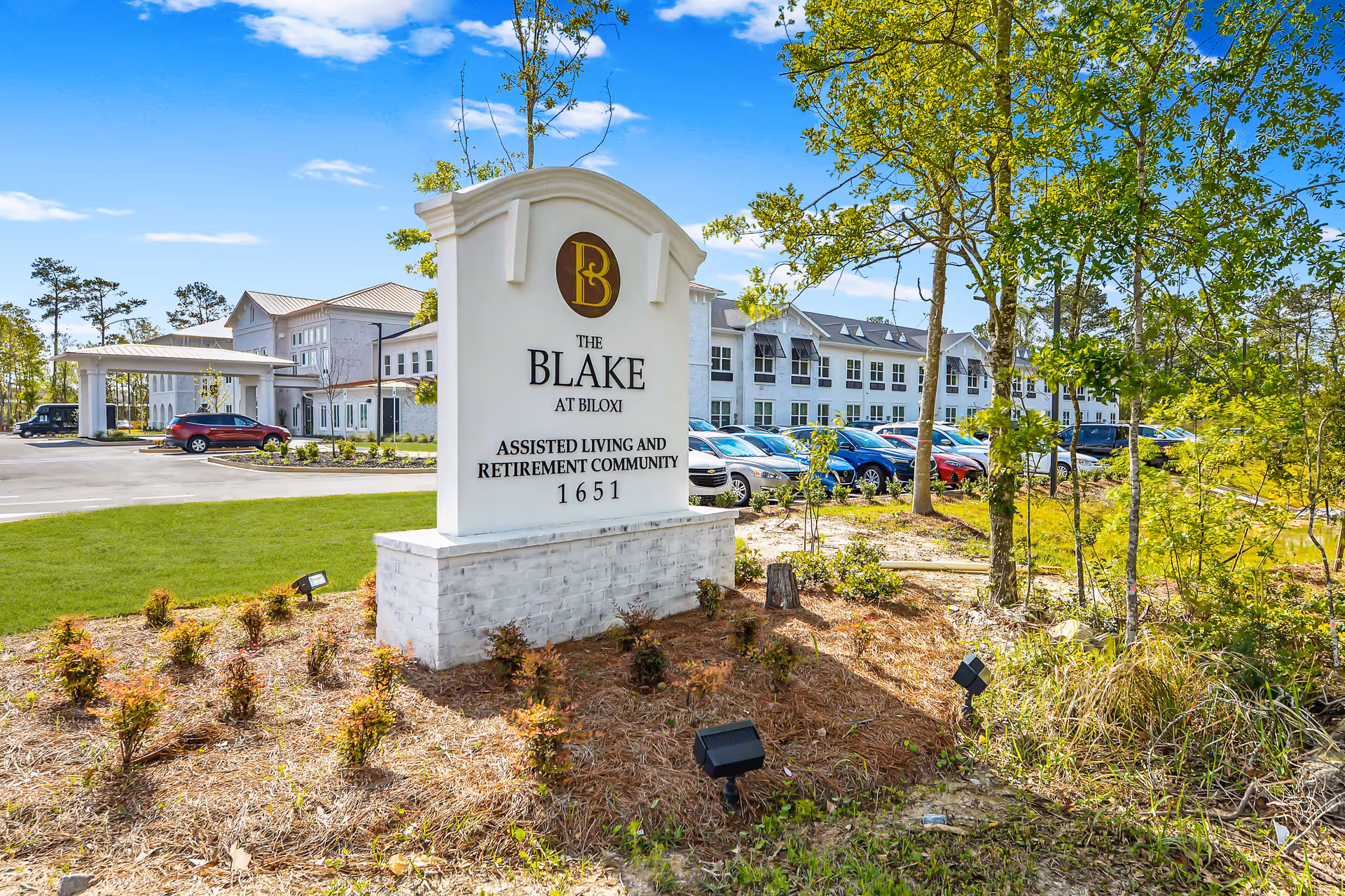 Outdoor view of The Blake at Biloxi assisted living and retirement community sign with the building and parking lot in the background under a blue sky.