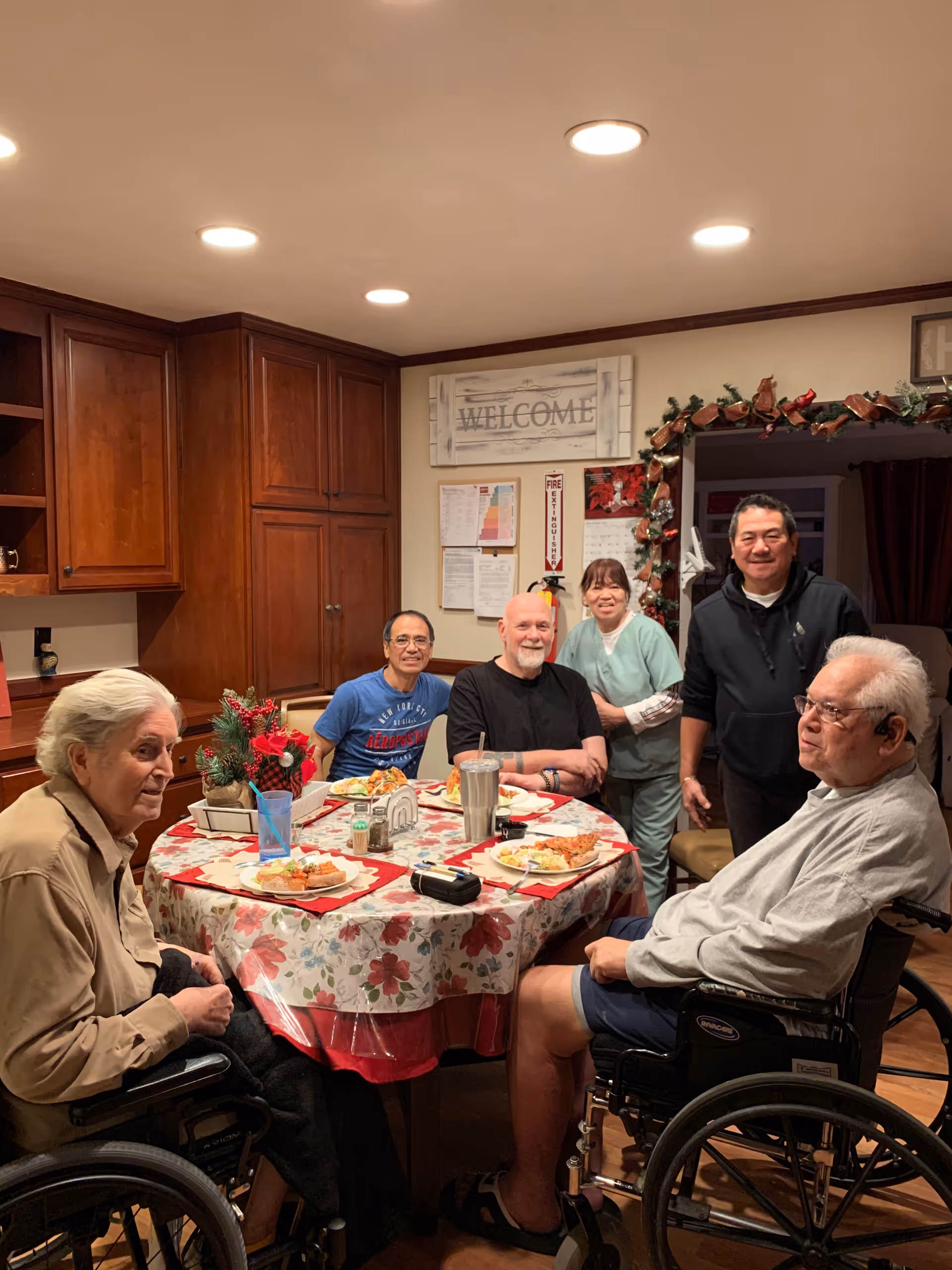 A group of residents and staff sit around a festively decorated dining table in a warmly lit room with a "WELCOME" sign on the wall.
