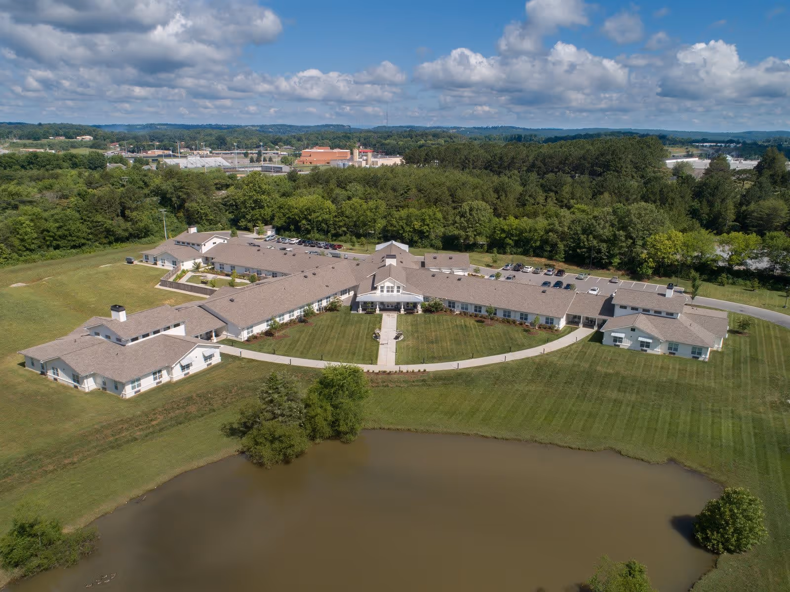 Aerial view of Dominion Senior Living of Athens facility showing a large, single-story building with multiple wings surrounded by green lawns, trees, and a pond in the foreground under a partly cloudy sky.