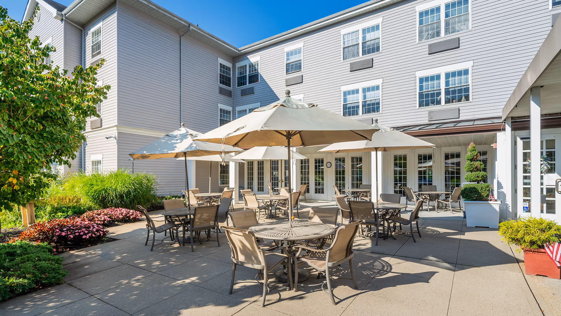 Outdoor patio area at Atria Hertlin Place with multiple round tables and chairs under large beige umbrellas, surrounded by greenery and flowers, adjacent to a multi-story building with many windows.
