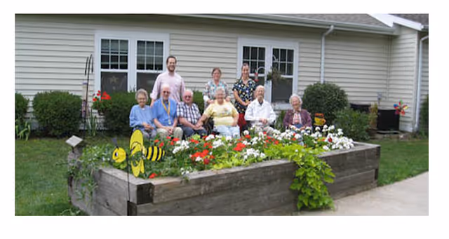 A group of elderly people and two caregivers sitting and standing behind a raised garden bed filled with colorful flowers outside a single-story building with beige siding and white-framed windows.