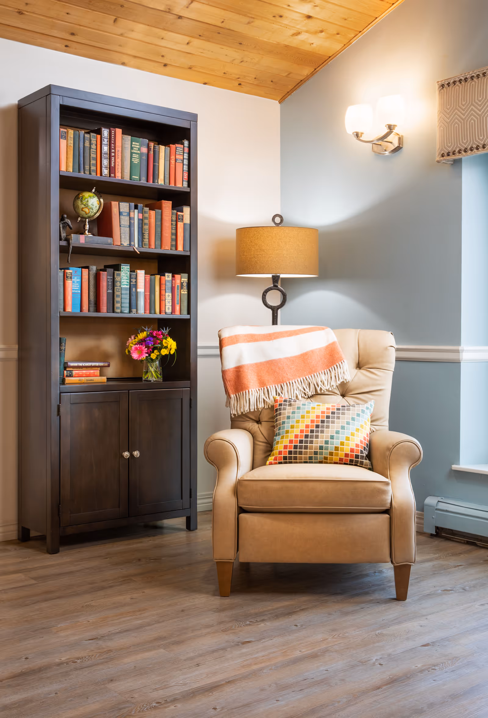 A cozy reading nook featuring a beige armchair with a colorful geometric pillow and an orange and white striped throw blanket draped over the back. Next to the chair is a dark wooden bookshelf filled with books and a small globe, with a vase of fresh flowers on one of the shelves. A floor lamp with a beige shade stands behind the chair, and a wall sconce with two lights is mounted on the light blue and white wall. The room has wooden flooring and a wooden ceiling.
