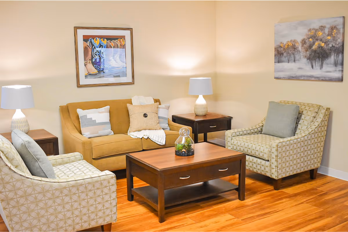 A cozy living room with a mustard yellow sofa adorned with three pillows and a throw blanket, two patterned armchairs with gray cushions, a wooden coffee table with a small decorative plant, two side tables each with a lamp, and two framed paintings on the beige walls.