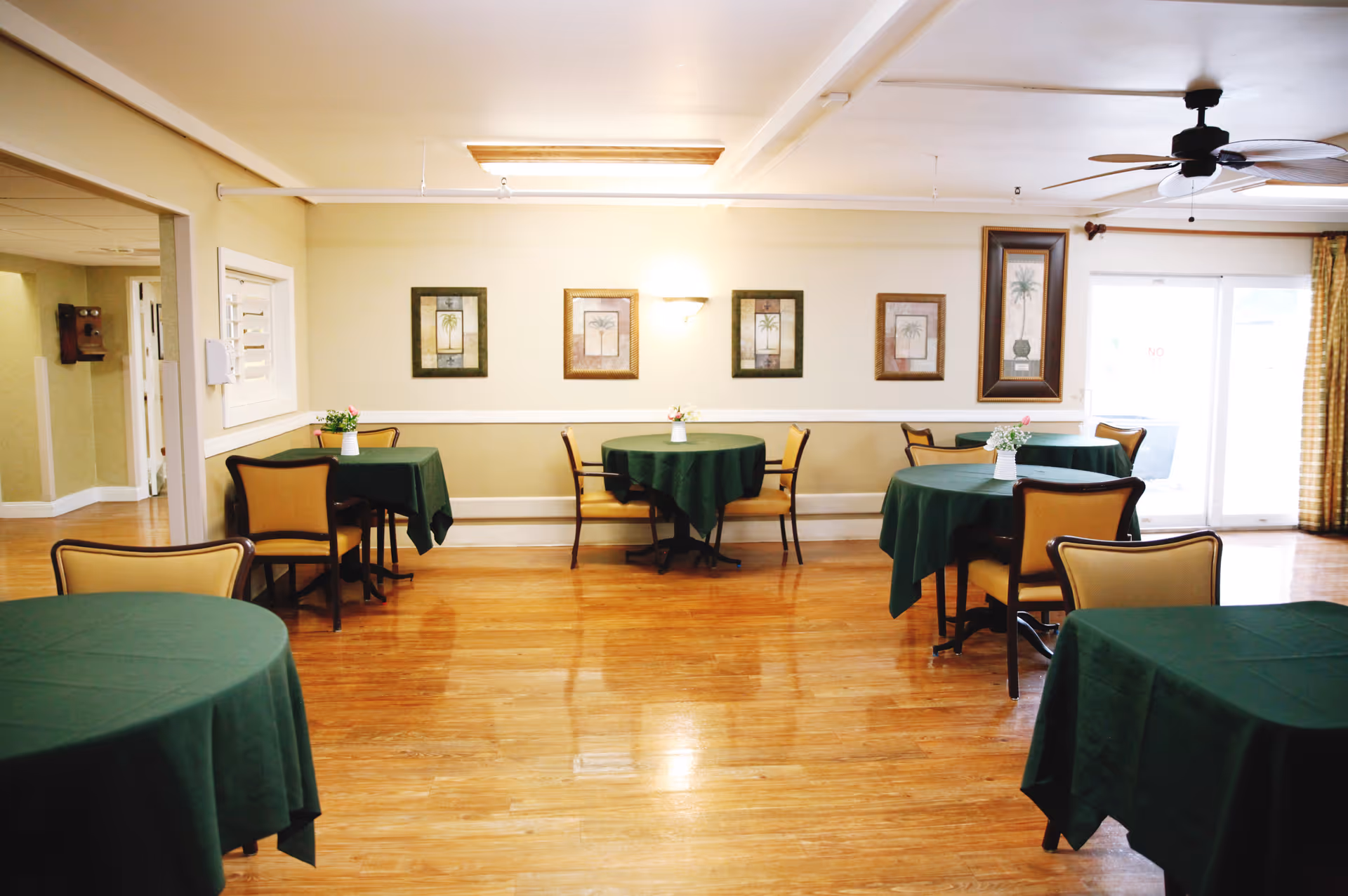 Dining room with multiple round tables draped in green tablecloths, chairs, hardwood floors, and framed artwork on the walls.