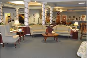 A spacious living room area with multiple beige upholstered sofas and armchairs arranged around wooden coffee tables. The room features several white columns wrapped with garlands, soft lighting from table lamps, and decorations on the walls. The carpet is blue, and the overall atmosphere is cozy and inviting.