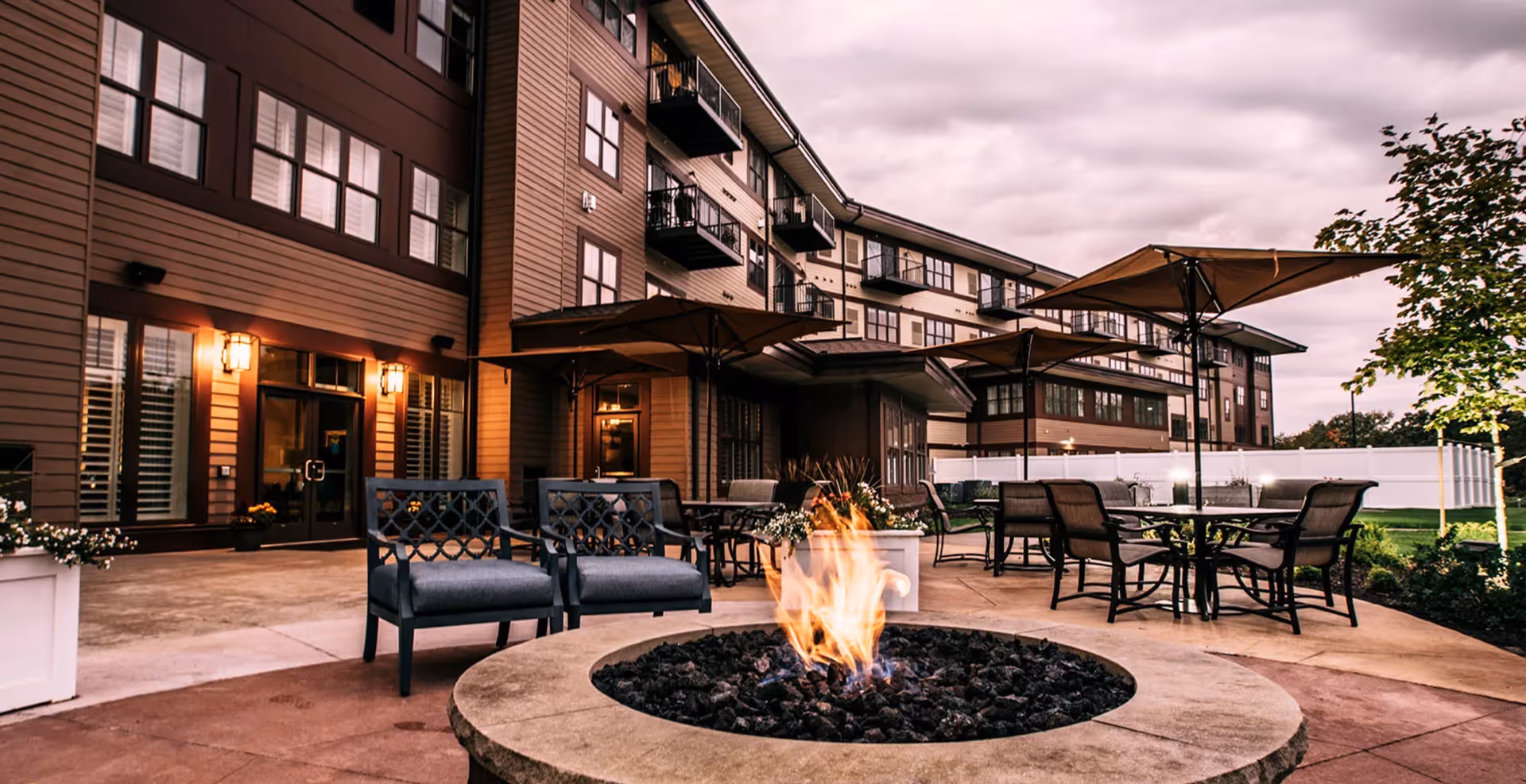 Outdoor patio at a senior living facility with a central fire pit, chairs, tables with umbrellas, and the building facade in the background.