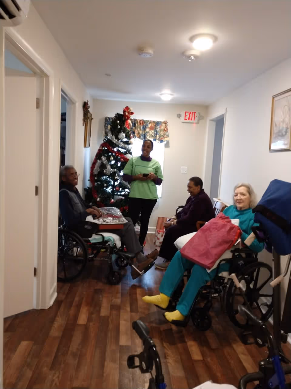 A hallway in Magnolia House Assisted Living decorated for Christmas with a Christmas tree. Three elderly residents, two in wheelchairs and one seated, are present along with a staff member standing and smiling. The floor is wooden, and there is an exit sign above a door at the end of the hallway.