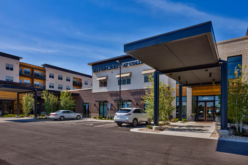 Exterior view of Silvercrest at College View Senior Living facility showing the main entrance with a covered drop-off area, parked cars, and a multi-story building under a clear blue sky.