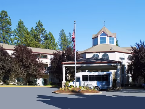 Front exterior of a multi-story senior living building with a covered porte-cochere, an American flag, and a shuttle van parked in front.