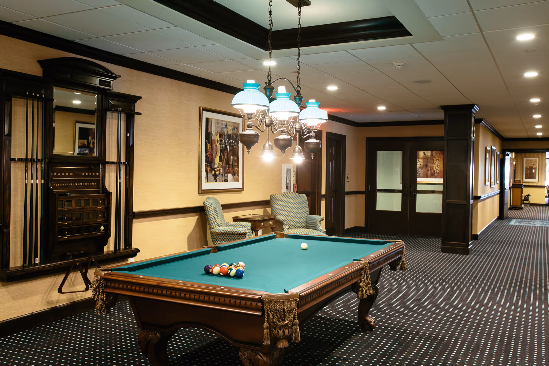 An indoor recreational area featuring a classic wooden pool table with billiard balls arranged for a game. Above the table hangs a vintage-style chandelier with multiple lamps. Behind the pool table, there are two upholstered armchairs and a small side table against a beige wall with framed artwork. The room has dark wood trim and a patterned carpet, with a hallway extending to the right.