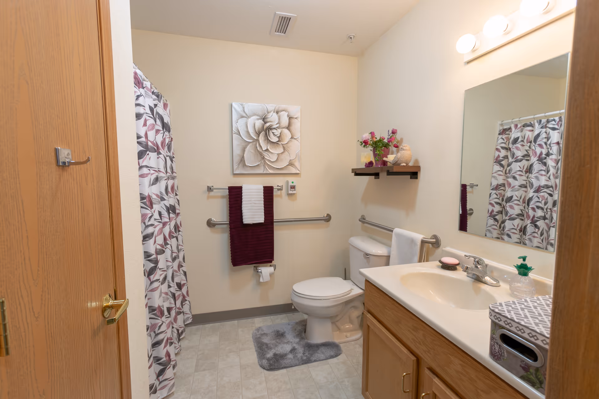A clean and well-lit bathroom with a toilet, sink, and shower. The shower has a curtain with a leaf pattern in shades of purple and gray. There are grab bars near the toilet and sink for support. A burgundy and white towel hangs on a bar above the toilet paper holder. Above the toilet is a small shelf with a vase of flowers and a teddy bear. A large mirror is mounted above the sink, which has a soap dispenser and a tissue box on the countertop. The walls are light beige, and the floor has light-colored tiles.