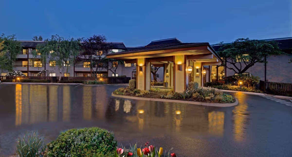 Evening view of the entrance to Madison House Assisted Living Community, featuring a covered drop-off area with warm lighting, surrounded by landscaped greenery and a multi-story building in the background with illuminated windows.