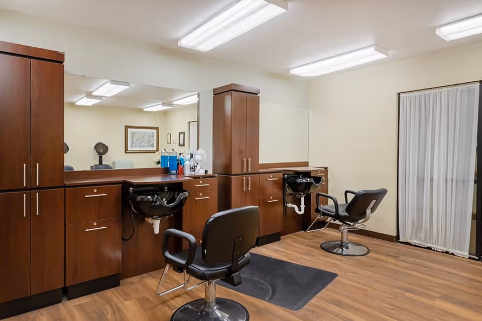 Interior view of a hair salon area with two black salon chairs in front of wooden cabinetry and black wash basins. A large mirror spans the wall behind the chairs, and the floor is wood with black mats under the chairs. Fluorescent lights illuminate the room, and a window with sheer white curtains is visible on the right.