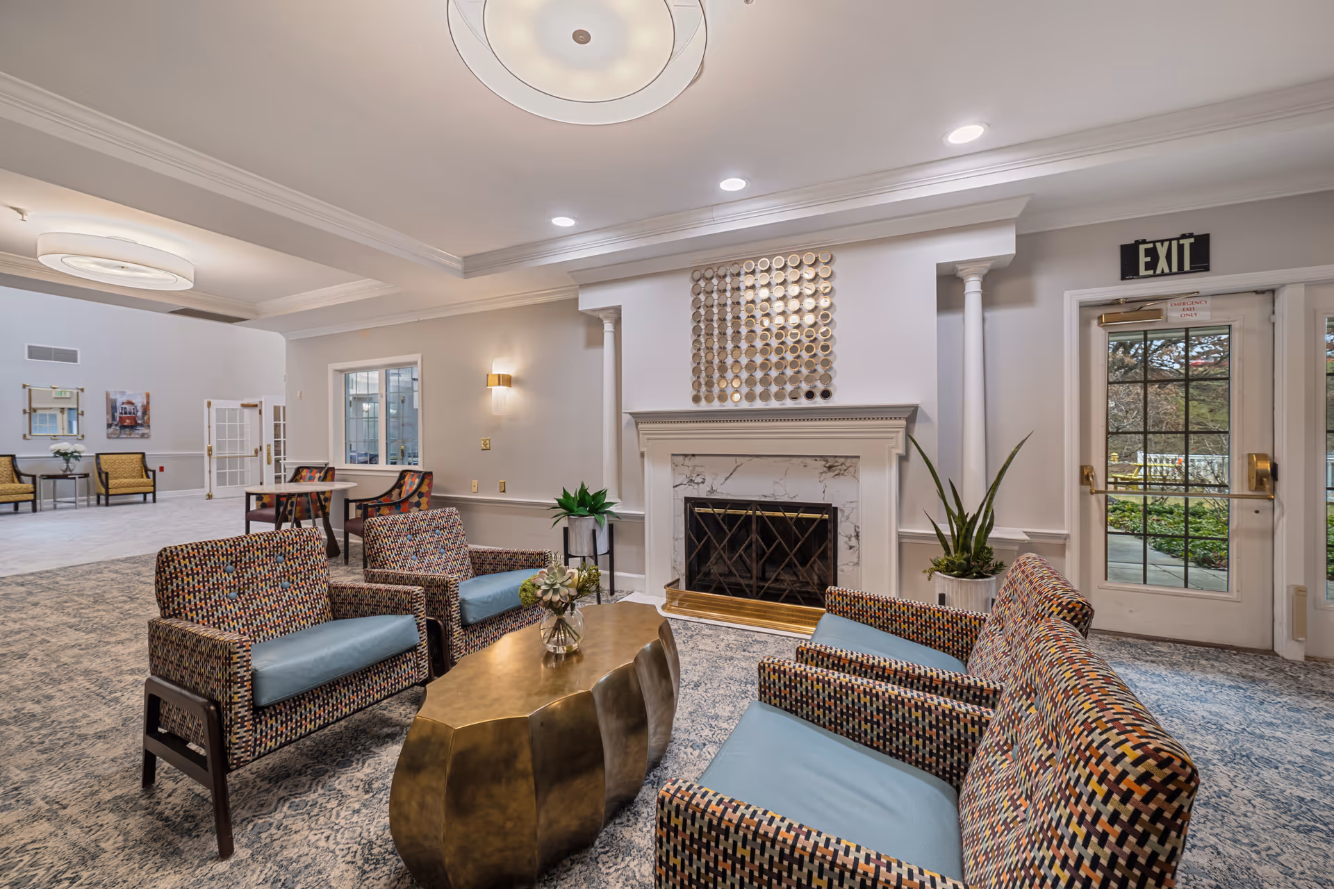 Common lounge with patterned armchairs and a brass coffee table facing a fireplace and exit doors.
