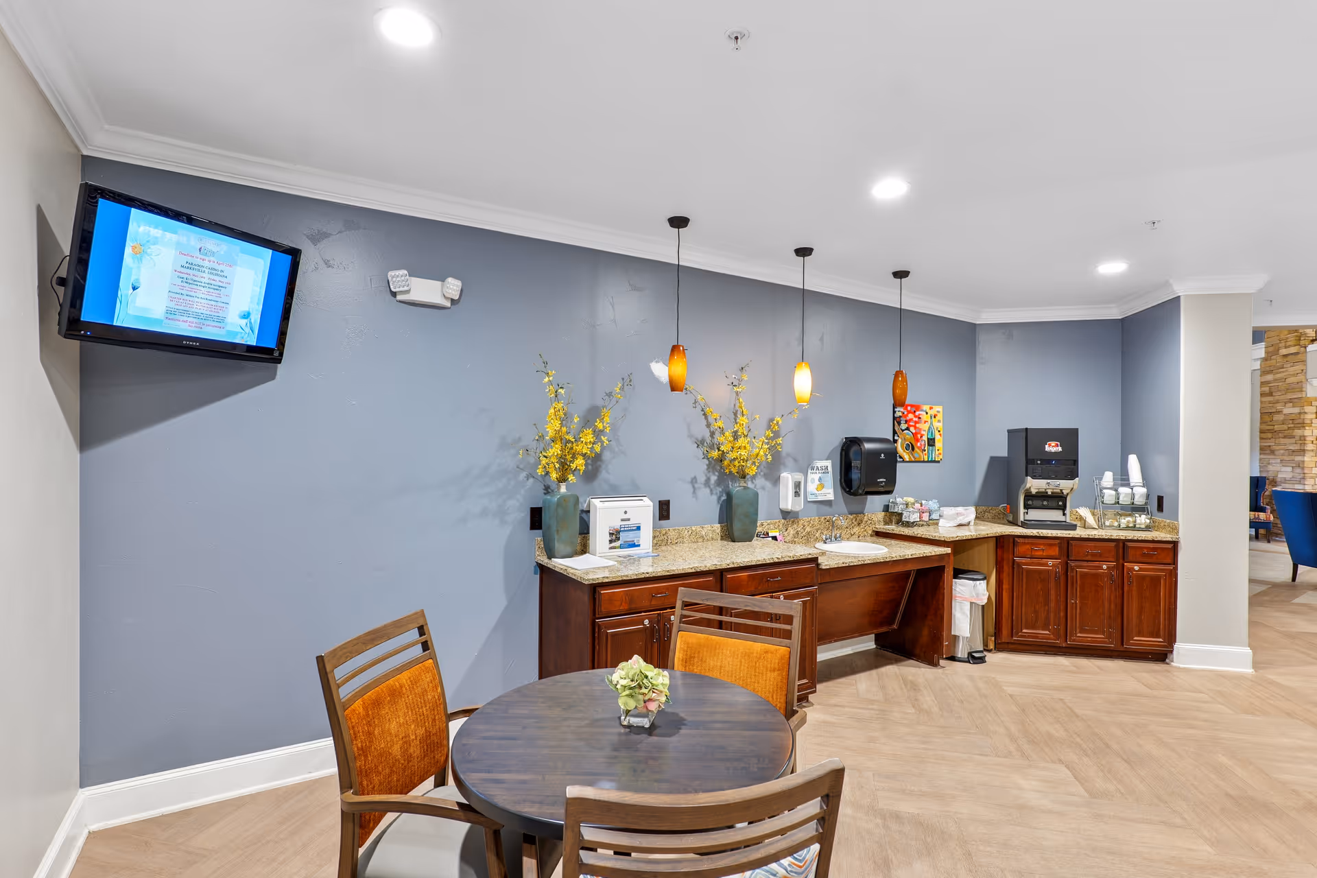 Common area with a round table and chairs in front of a countertop coffee and sink station with pendant lights and a wall-mounted TV.