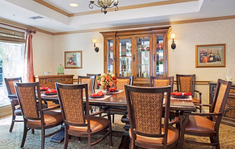 A well-lit dining room with a large wooden dining table surrounded by eight upholstered chairs. The table is set with red napkins and placemats, and a floral centerpiece. Behind the table is a wooden china cabinet displaying glassware and decorative items. The room has beige wallpaper, framed artwork on the walls, a window with curtains, and a chandelier hanging from a tray ceiling.