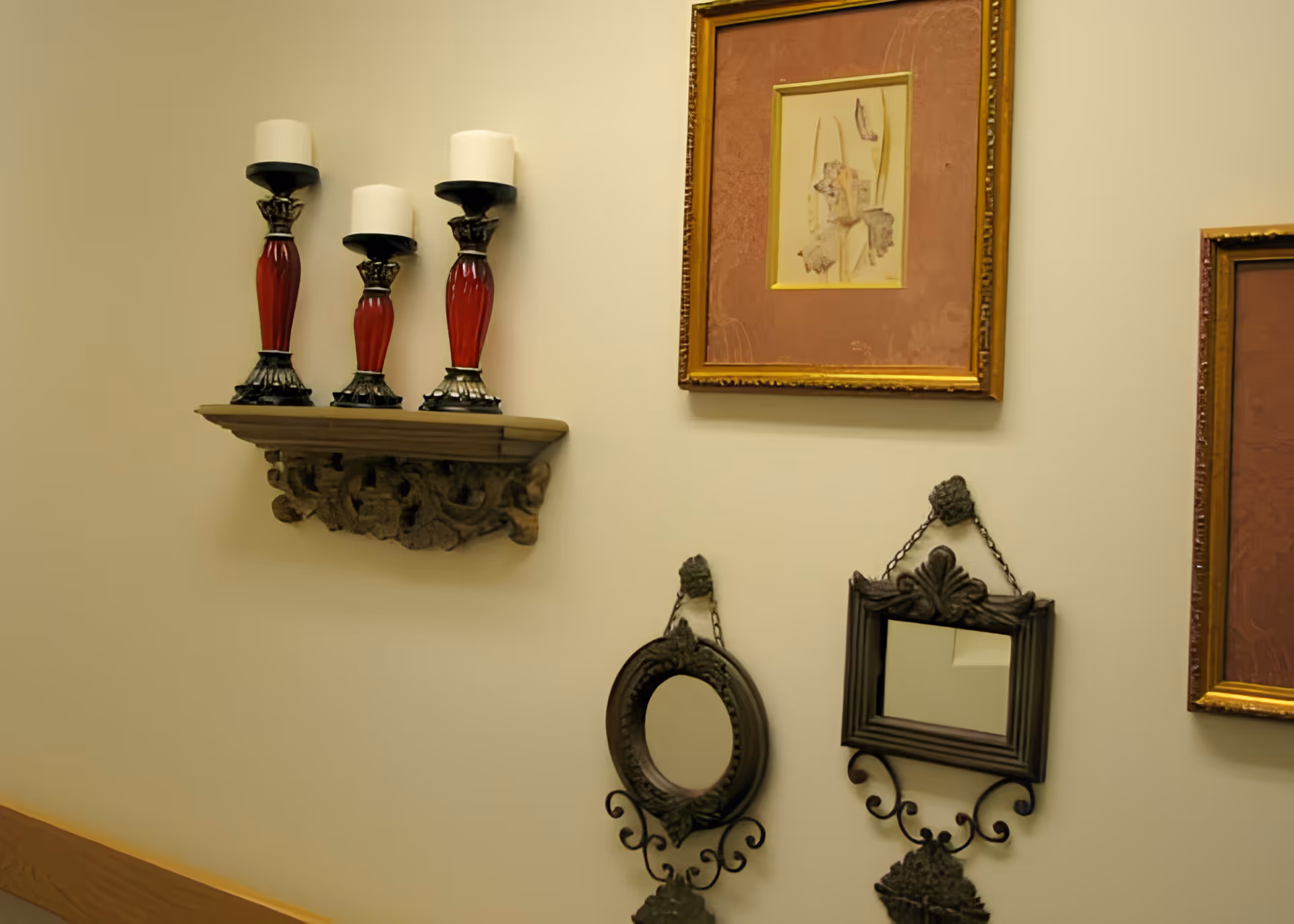 A beige wall decorated with a small wooden shelf holding three red and black candle holders with white candles. Below the shelf are two ornate black metal-framed mirrors, one oval and one rectangular, hanging on the wall. To the right, there are two framed artworks with gold frames and reddish mats.