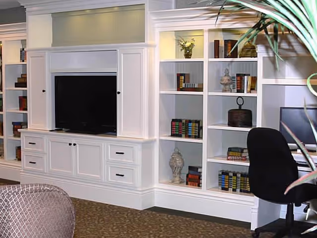 A cozy interior space featuring a white built-in entertainment center with a flat-screen TV, surrounded by shelves filled with decorative books and ornaments. To the right, there is a black office chair in front of a desk with a computer monitor. A plant partially frames the right side of the image.