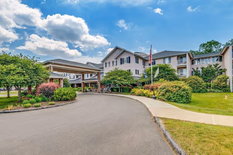 Front entrance and covered porte-cochere of a multi-story senior living building with a driveway, landscaped greenery, and an American flag under a partly cloudy sky.