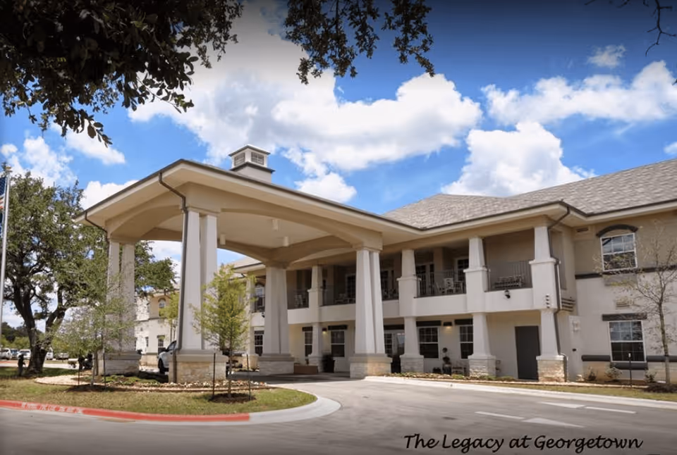 Exterior view of a two-story senior living facility building with a covered entrance supported by large columns. The building has multiple windows and a balcony area on the second floor. Trees and landscaping surround the driveway, and the sky is partly cloudy with blue patches.