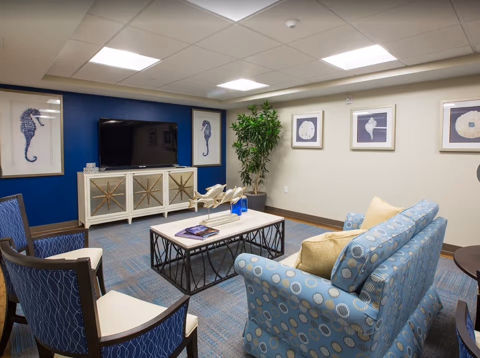 Well-lit cozy lounge with blue-accented walls, patterned sofas and chairs facing a TV on a decorative console and a central coffee table.