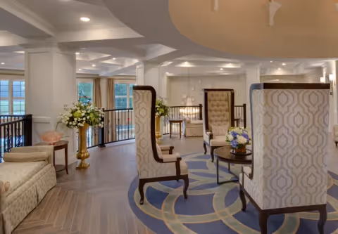 A spacious and elegant common area with high-back patterned armchairs arranged around a circular rug with a geometric design. The room features large windows with views of greenery outside, decorative flower arrangements in tall vases, and soft lighting from recessed ceiling lights.