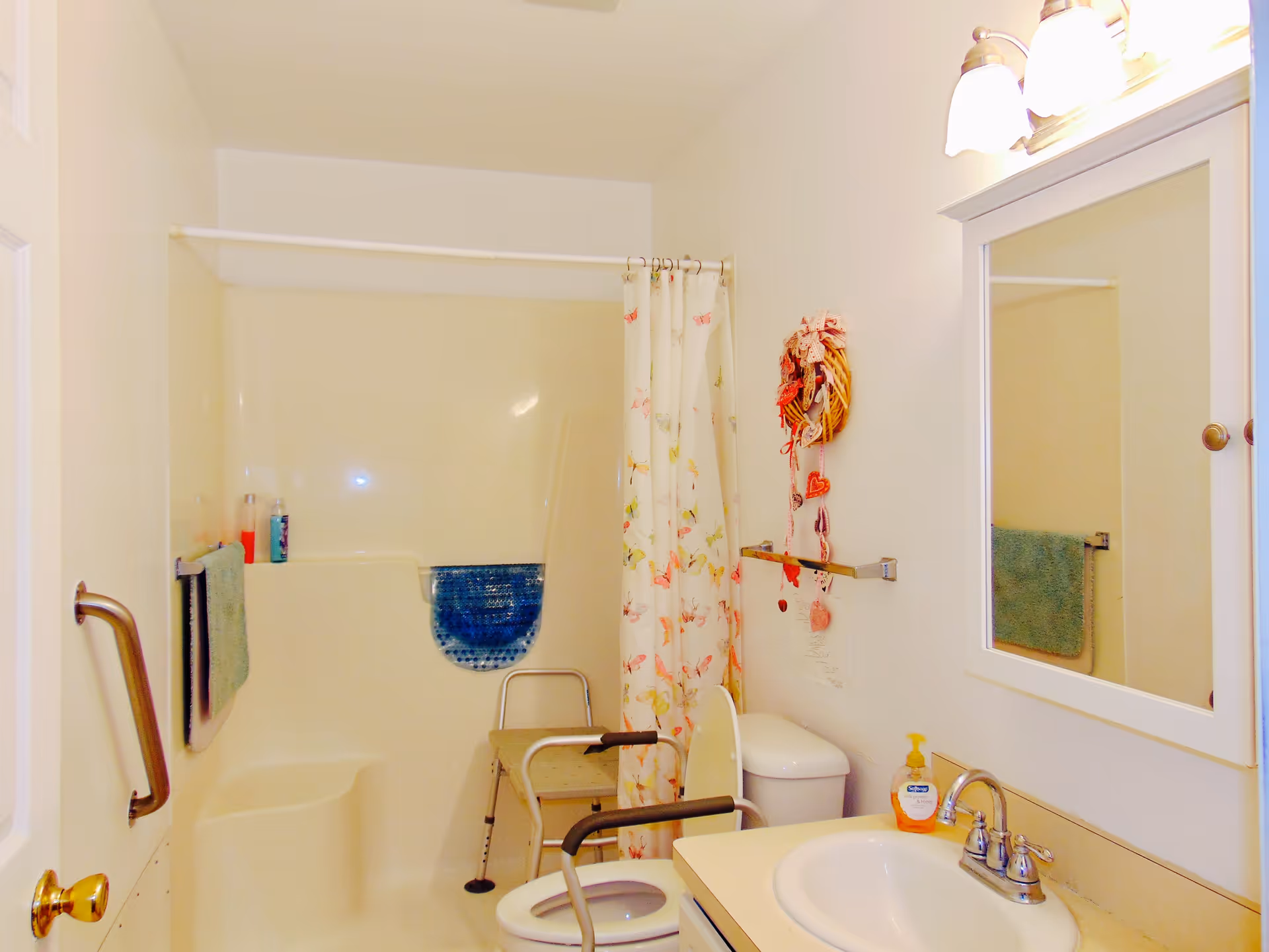 A small bathroom with a shower and bathtub combination, a floral shower curtain, a toilet with a raised seat and arm supports, a sink with a mirror cabinet above it, a towel rack with a green towel, and a grab bar on the wall near the door. There is also a decorative hanging on the wall above the toilet.