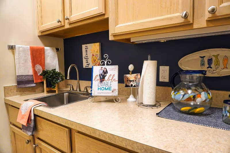 A kitchen countertop with a stainless steel sink, a faucet, and wooden cabinets above and below. On the counter, there is a decorative book stand holding a book titled 'lovable LIVABLE HOME,' a paper towel holder with paper towels, a small photo frame with a cat wearing glasses, and a glass pitcher with a blue lid and colorful fish design. A towel rack on the left wall holds two towels, one orange and one white with a shell design. The wall behind the counter is painted dark blue with a small decorative sign that says 'welcome' and a fish-shaped wall decoration.