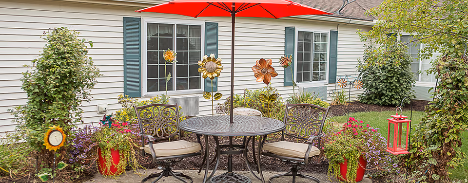 Outdoor patio area with a round metal table and two cushioned chairs under a red umbrella, surrounded by potted plants, decorative metal flowers, and greenery in front of a building with white siding and green shutters.