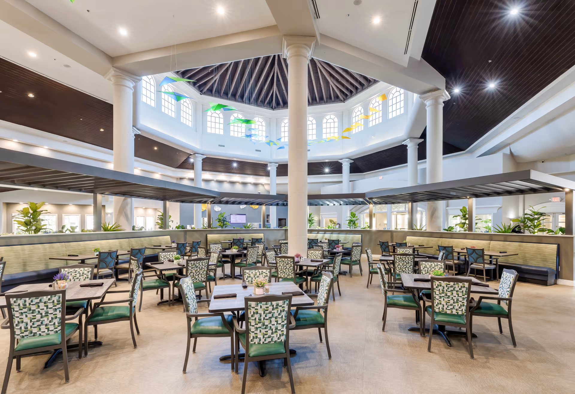 A spacious and well-lit dining room with multiple tables and chairs arranged neatly. The room features a high ceiling with large windows near the top, allowing natural light to fill the space. There are decorative hanging elements in green and yellow colors near the ceiling. The walls have large columns and there are plants placed around the room, creating a welcoming atmosphere.