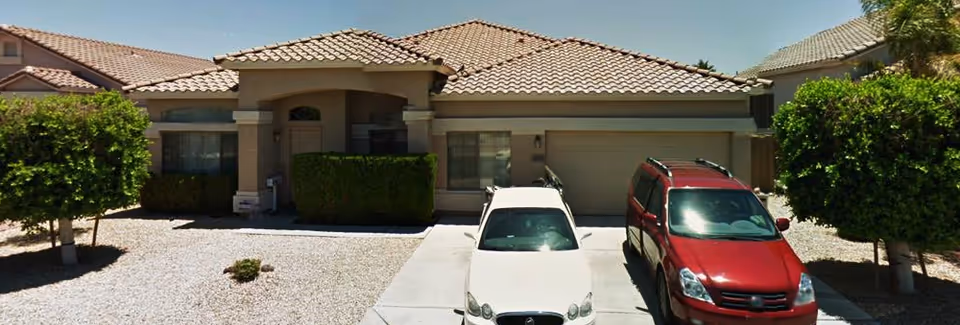 Front exterior view of a single-story house with a tiled roof, two cars parked in the driveway, and small trees and bushes in the front yard covered with gravel.