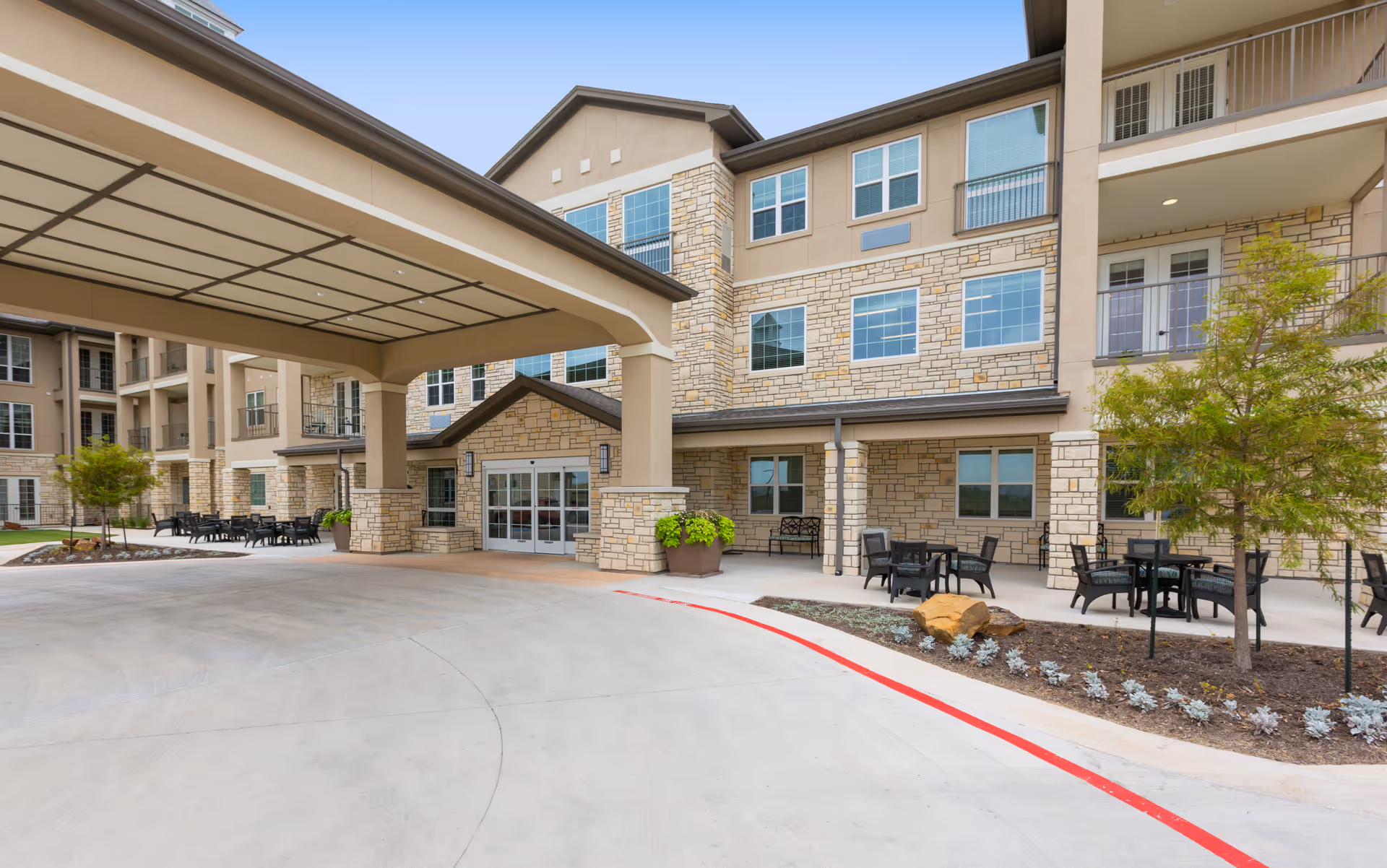Covered porte-cochere and entrance of a multi-story stone-faced senior living building with outdoor seating and landscaping.