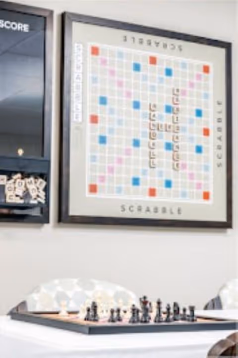 A framed Scrabble board mounted on a wall above a table with a chess set arranged for play. The setting appears to be a recreational or common area with patterned chairs partially visible.